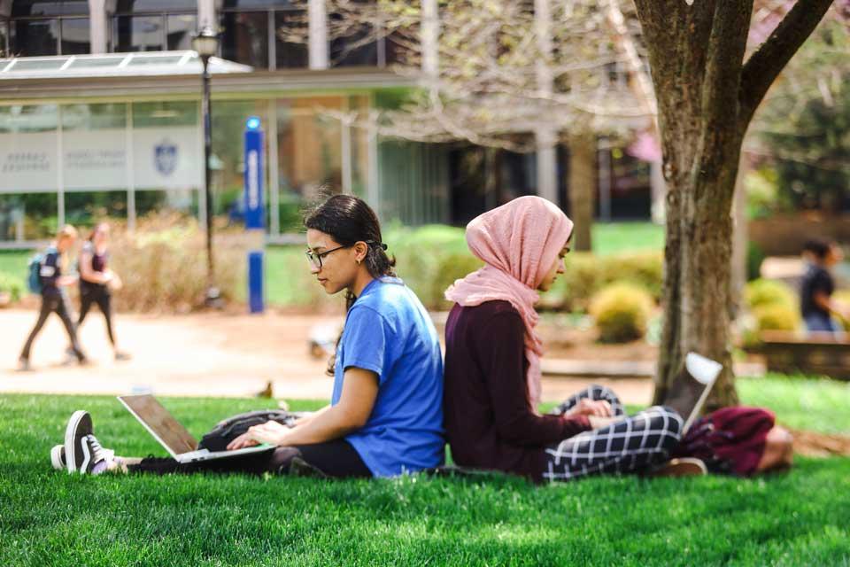 two students, one wearing a headscarf, sit back to back in the grass under a tree